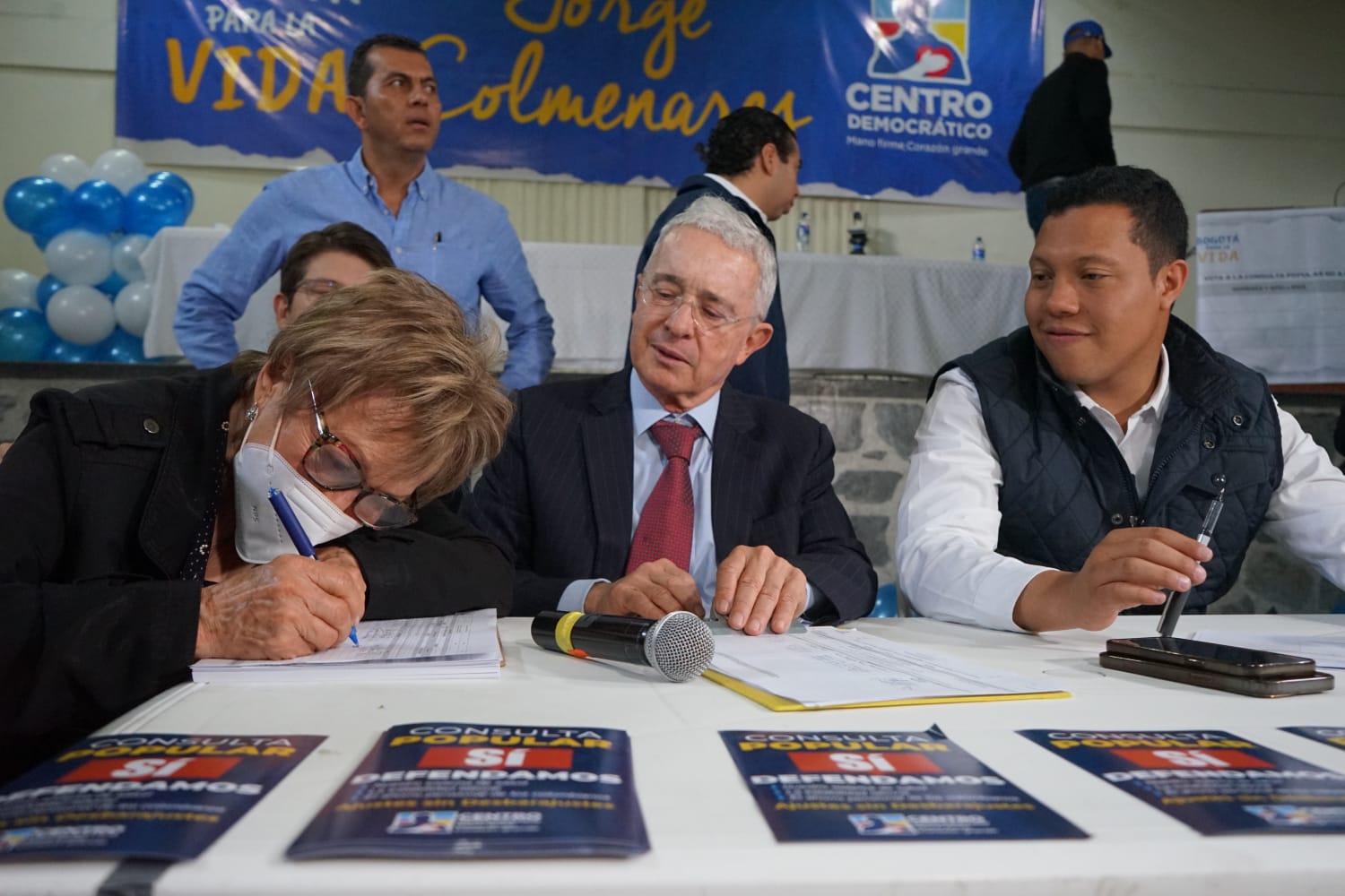 Foto en la que aparecen una ciudadana firmando junto a Alvaro Uribe y el concejal Colmenares