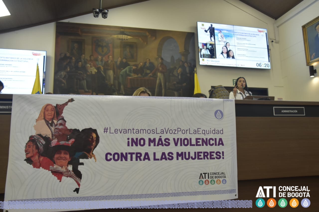 Fotografía en la que aparece la concejal ATi Quigua en el Atril del Recinto los Comuneros, junto a un pendón con un escrito que dice "No más violencia contra las mujeres
